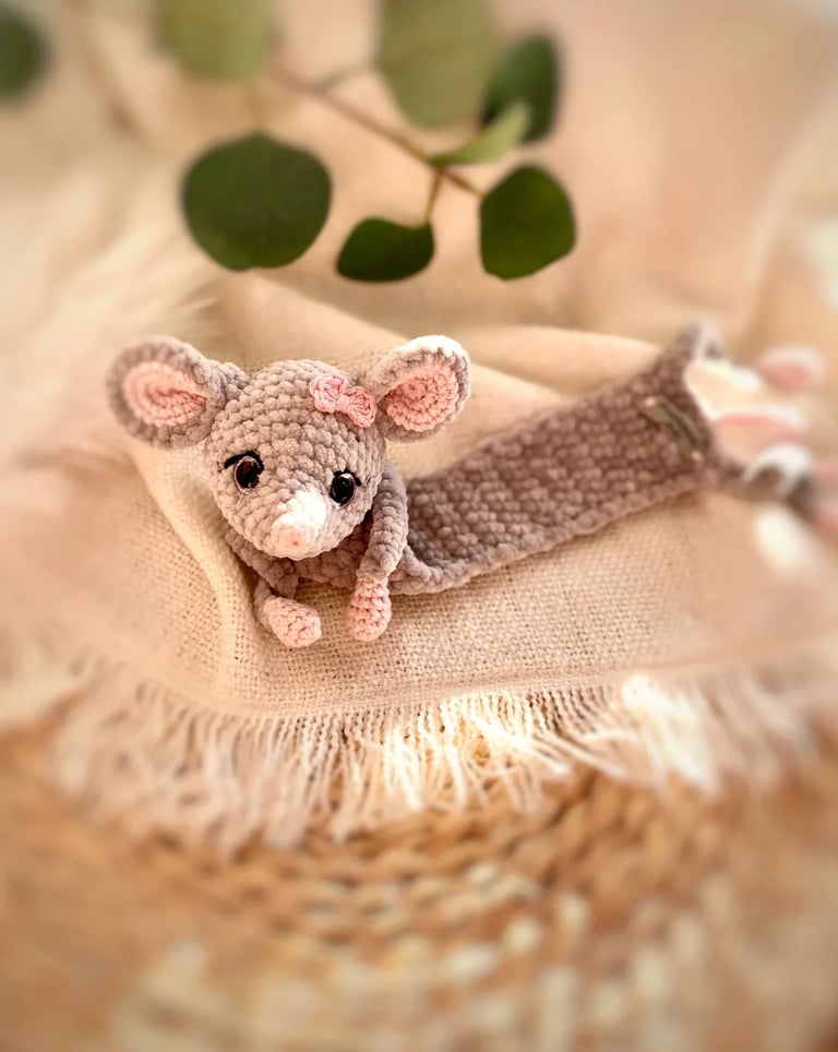 Small crocheted pink mouse toy with black beads for eyes, sitting on a woven beige fabric with green leaves in the background