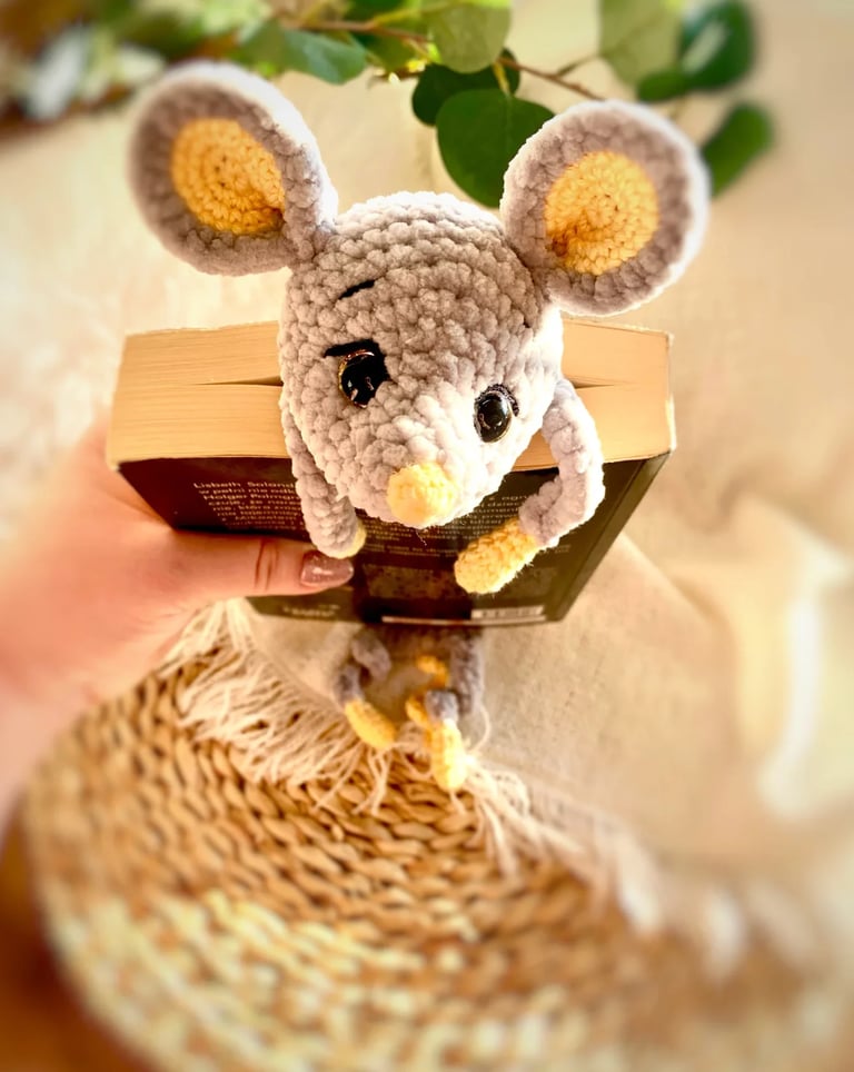 Crocheted beige and yellow mouse amigurumi sitting on a brown cardboard box with green foliage in background